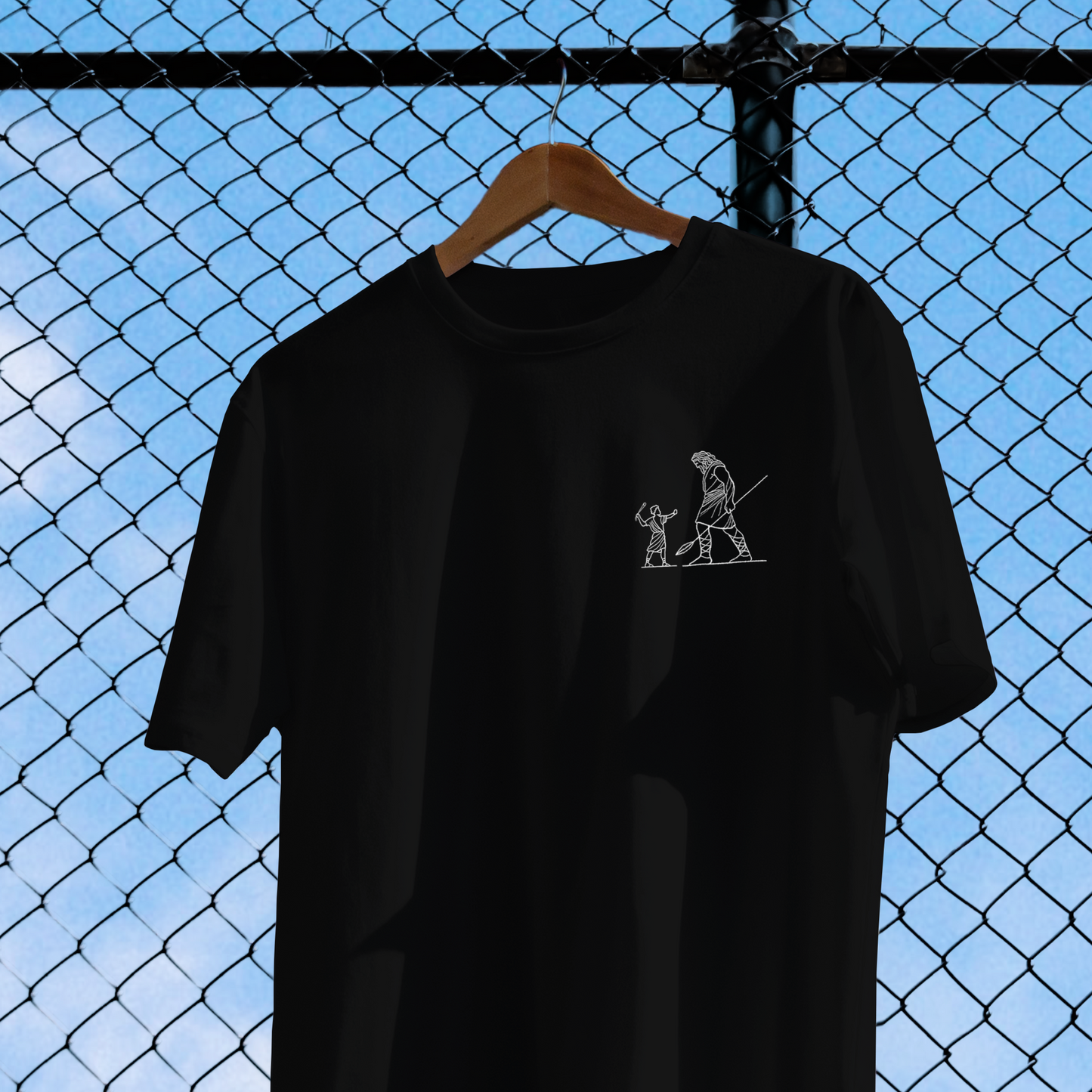 Black t-shirt with a small white graphic on a hanger against a chain-link fence background.