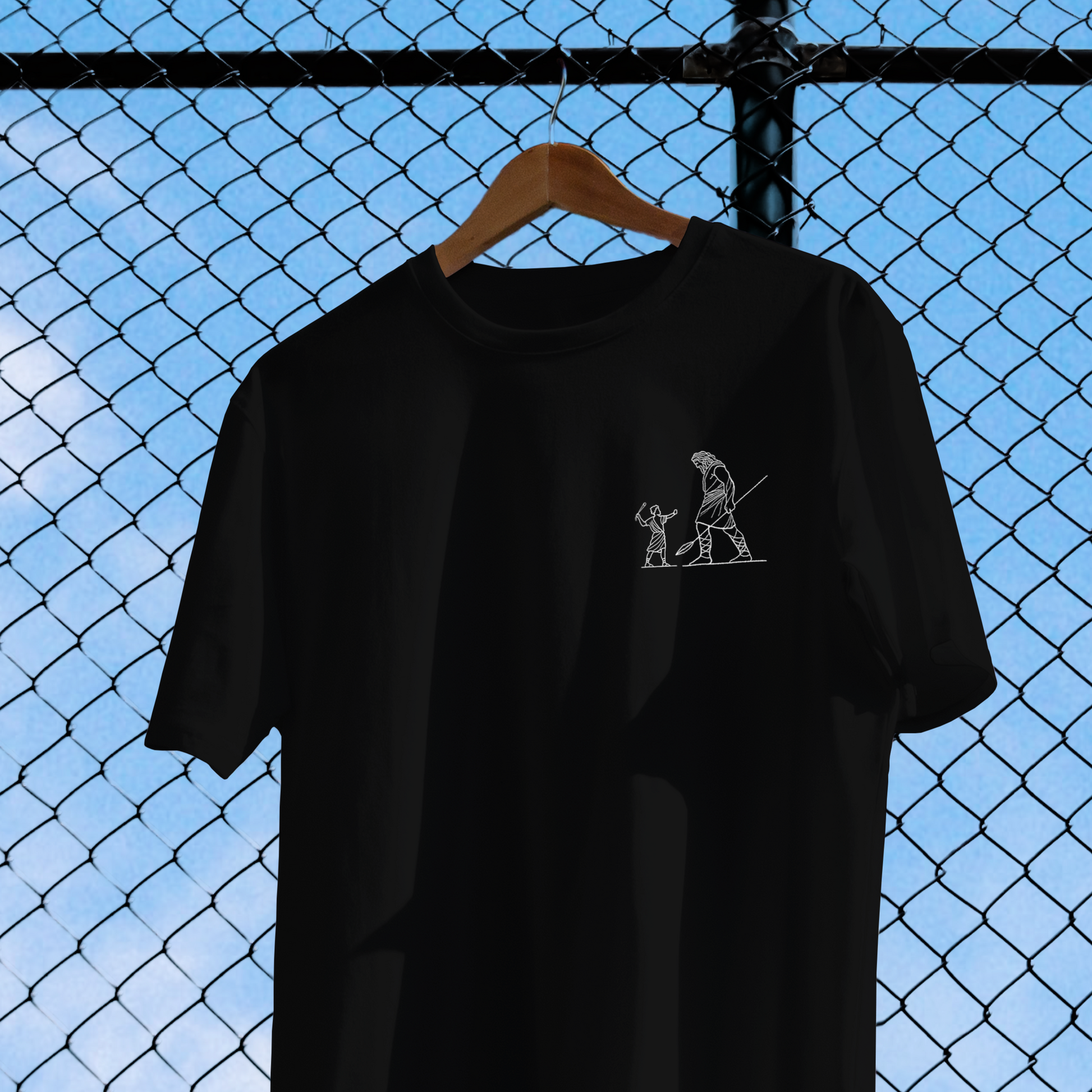 Black t-shirt with a small white graphic on a hanger against a chain-link fence background.