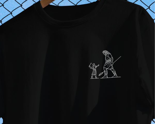Black t-shirt with a small white graphic on a hanger against a chain-link fence background.